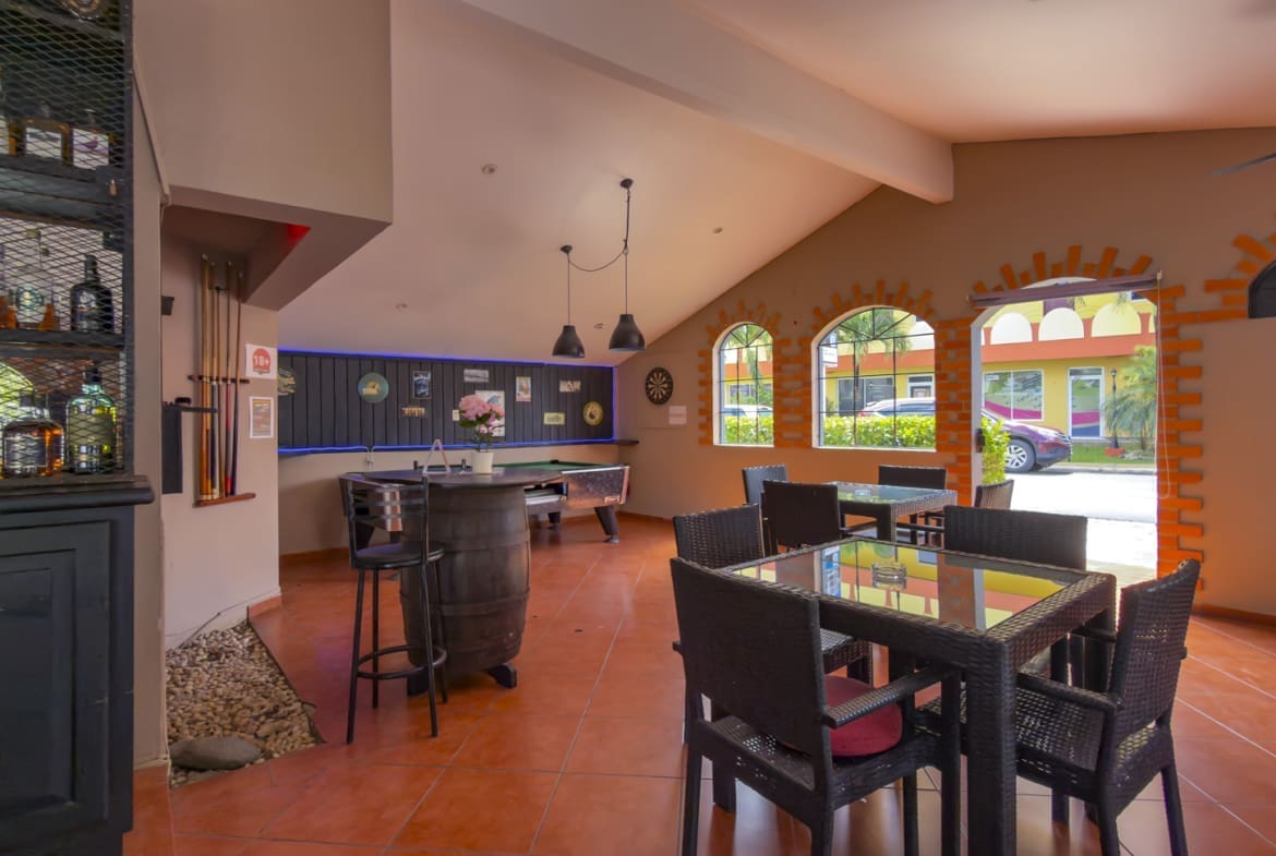 Indoor lounge with pool tables, bar, and wicker dining set under warm lighting; brick arches and large windows overlook outdoor seating area
