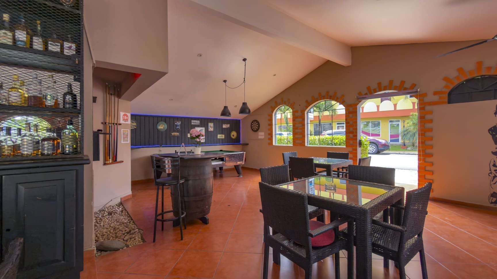 Indoor lounge with pool tables, bar, and wicker dining set under warm lighting; brick arches and large windows overlook outdoor seating area