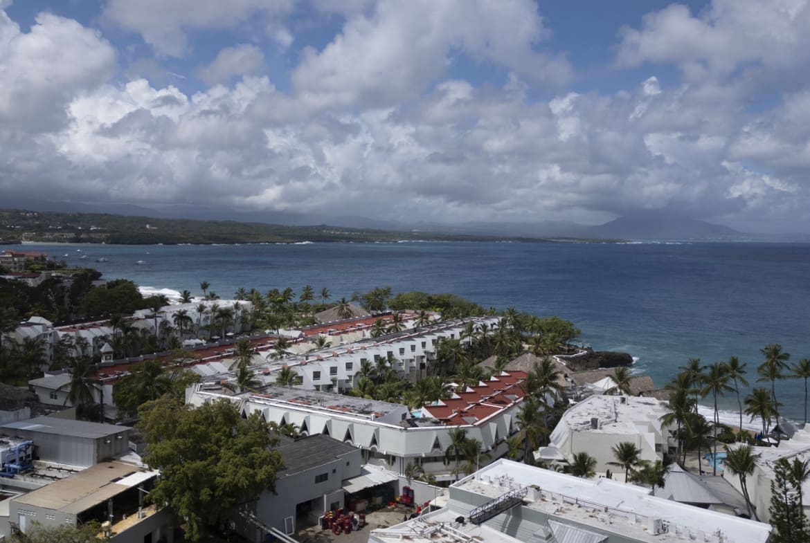 buyDRproperty Aerial view of a coastal resort with white buildings and palm trees overlooking the ocean. Sosua Real Estate