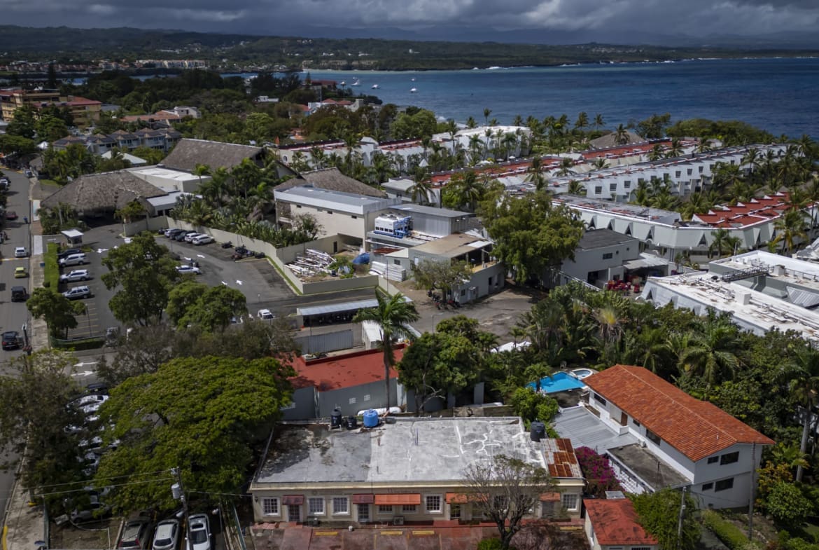 buyDRproperty Aerial view of a coastal town with buildings, roads, and the ocean under a cloudy sky. Sosua Real Estate