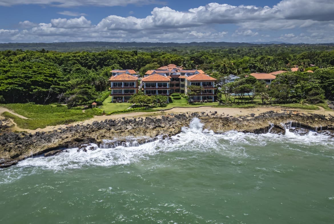 buyDRproperty Oceanfront villas with orange roofs on a rocky coastline, lush green trees, and blue sky. Sosua Real Estate