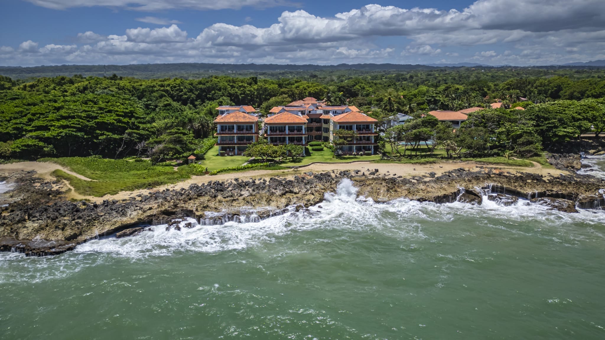 buyDRproperty Oceanfront villas with orange roofs on a rocky coastline, lush green trees, and blue sky. Sosua Real Estate