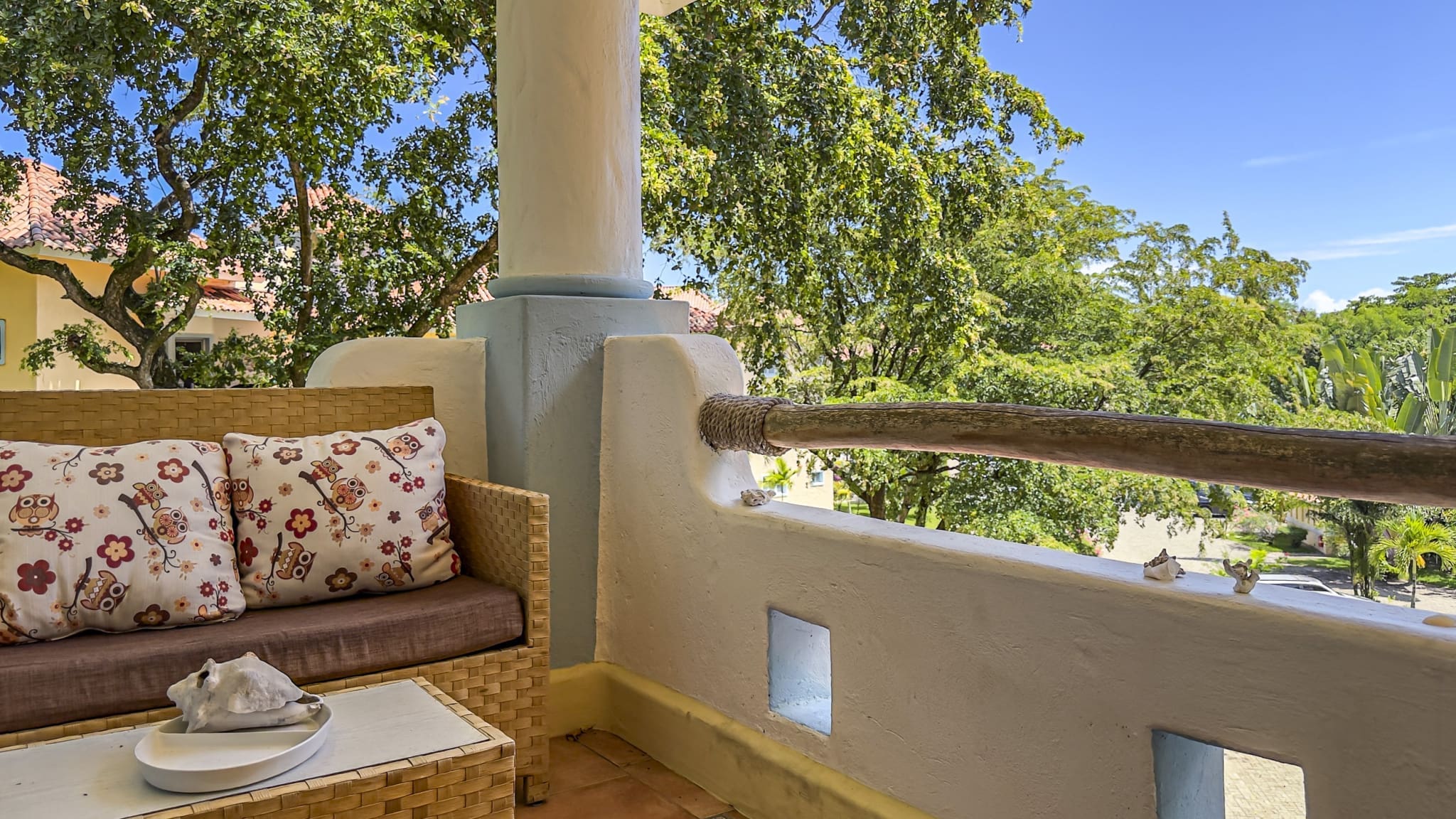 buyDRproperty Wicker sofa with owl pillows on a balcony with lush green trees and blue sky. Sosua Real Estate