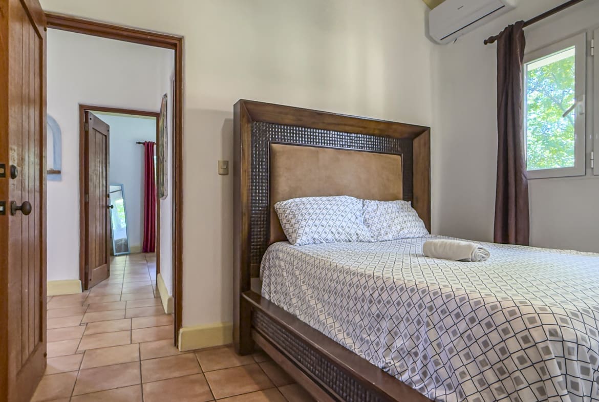 A large wooden bed with a padded headboard and geometric quilt in a sunlit bedroom; a rolled towel rests on the bed.