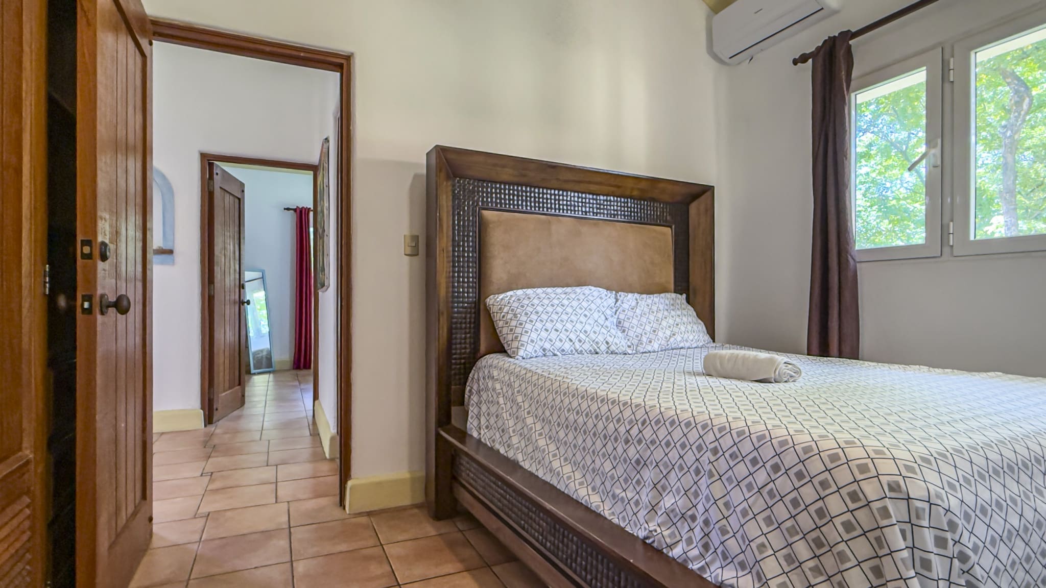 A large wooden bed with a padded headboard and geometric quilt in a sunlit bedroom; a rolled towel rests on the bed.
