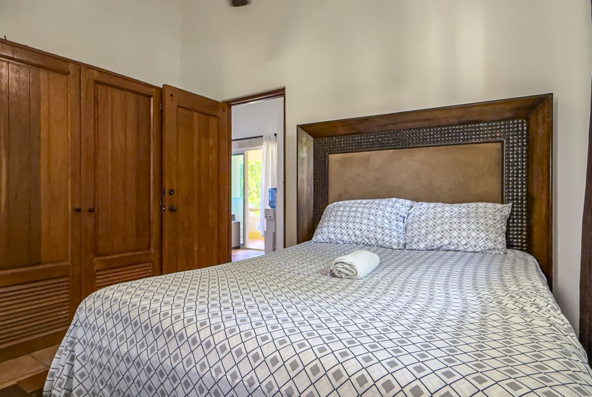 Bedroom with a large patterned bedspread, two pillows, and a rolled towel on the bed; wooden wardrobe on the left and an open doorway to a sunlit room.