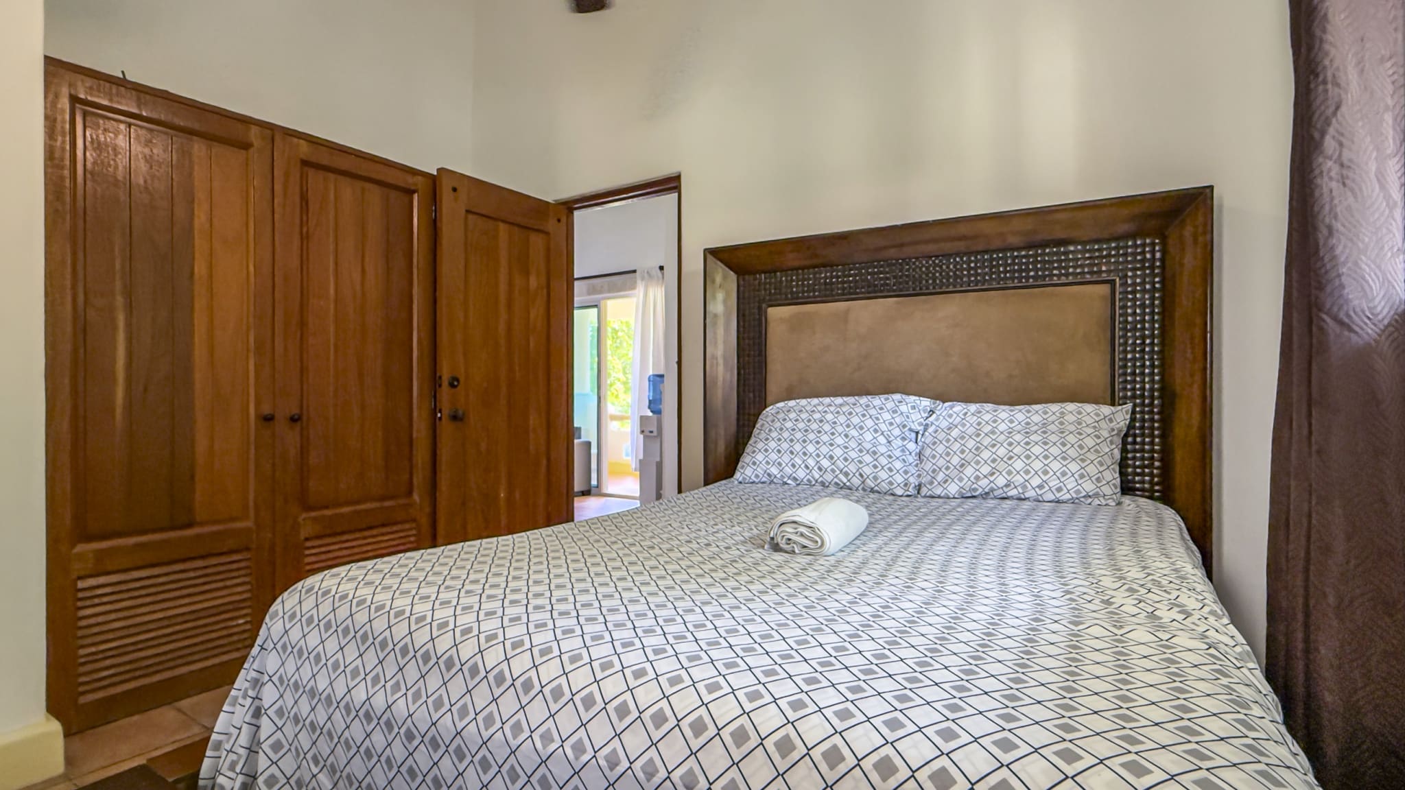 Bedroom with a large patterned bedspread, two pillows, and a rolled towel on the bed; wooden wardrobe on the left and an open doorway to a sunlit room.