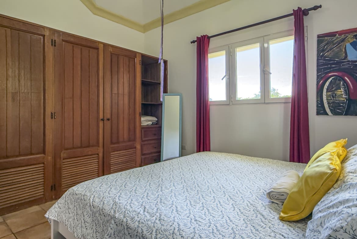 buyDRproperty Bedroom with wooden wardrobes, bed with yellow pillows, and a window with red curtains. Sosua Real Estate
