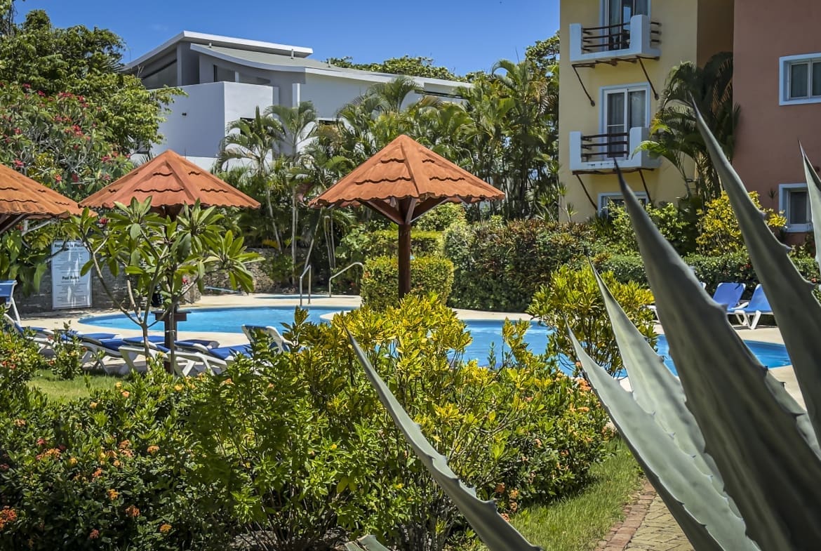 Poolside at a tropical resort with orange thatched umbrellas, lounge chairs, and lush greenery around a bright blue pool.