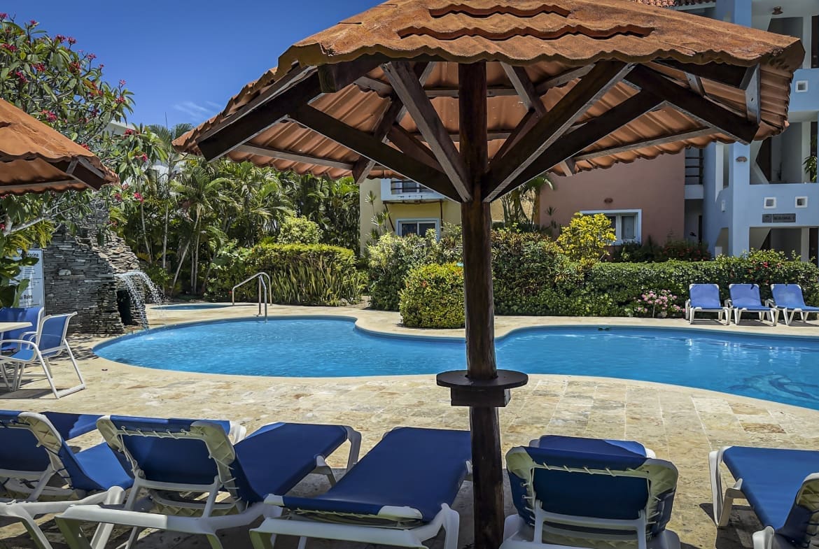Resort pool area with blue lounge chairs under large terracotta shade umbrellas, surrounded by tropical plants and pastel buildings beyond.