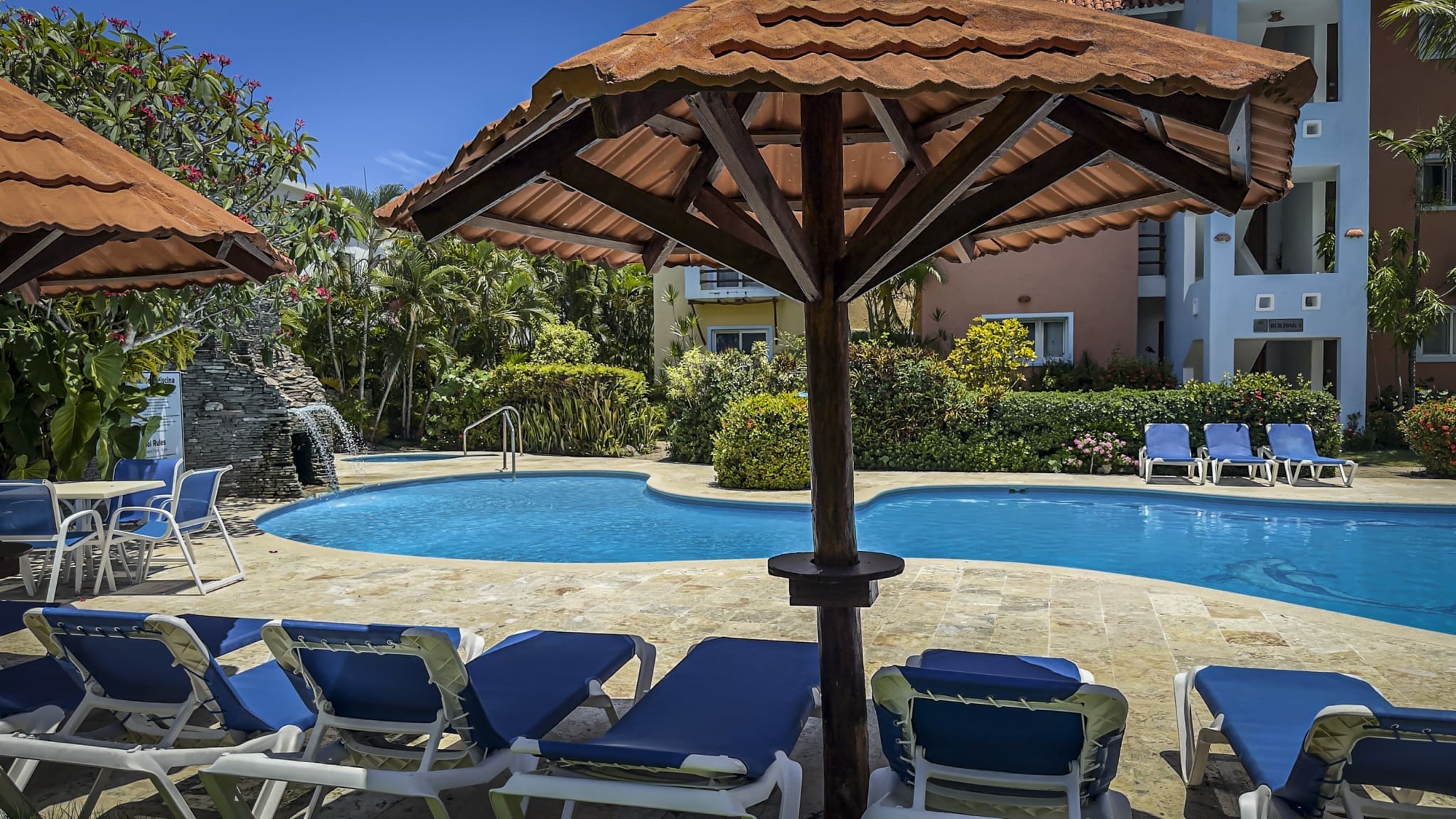 Resort pool area with blue lounge chairs under large terracotta shade umbrellas, surrounded by tropical plants and pastel buildings beyond.