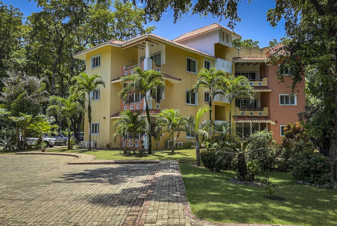 Colorful, multi-story apartment building set in a tropical garden with palm trees and a brick driveway beneath a blue sky.