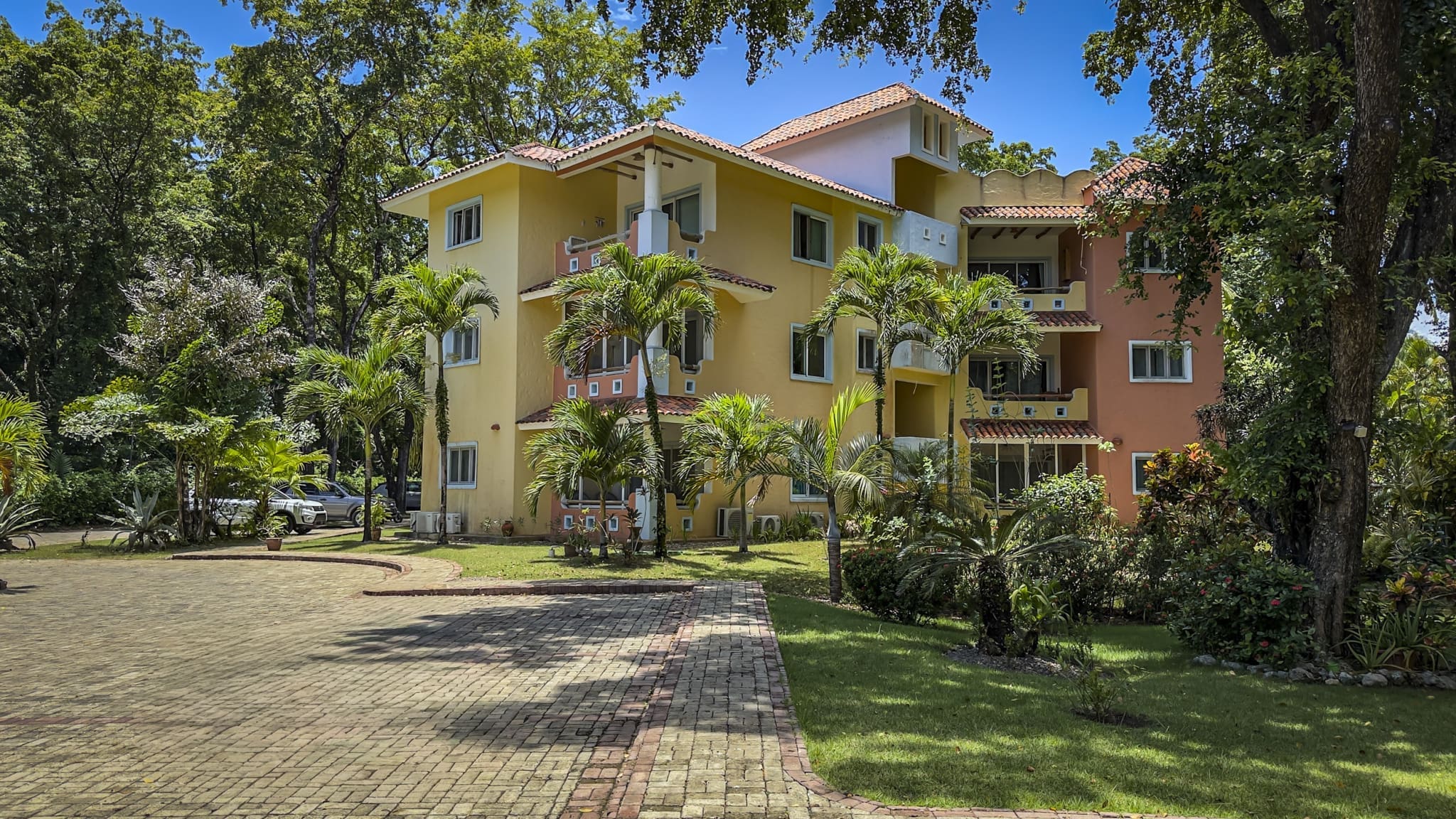 Colorful, multi-story apartment building set in a tropical garden with palm trees and a brick driveway beneath a blue sky.