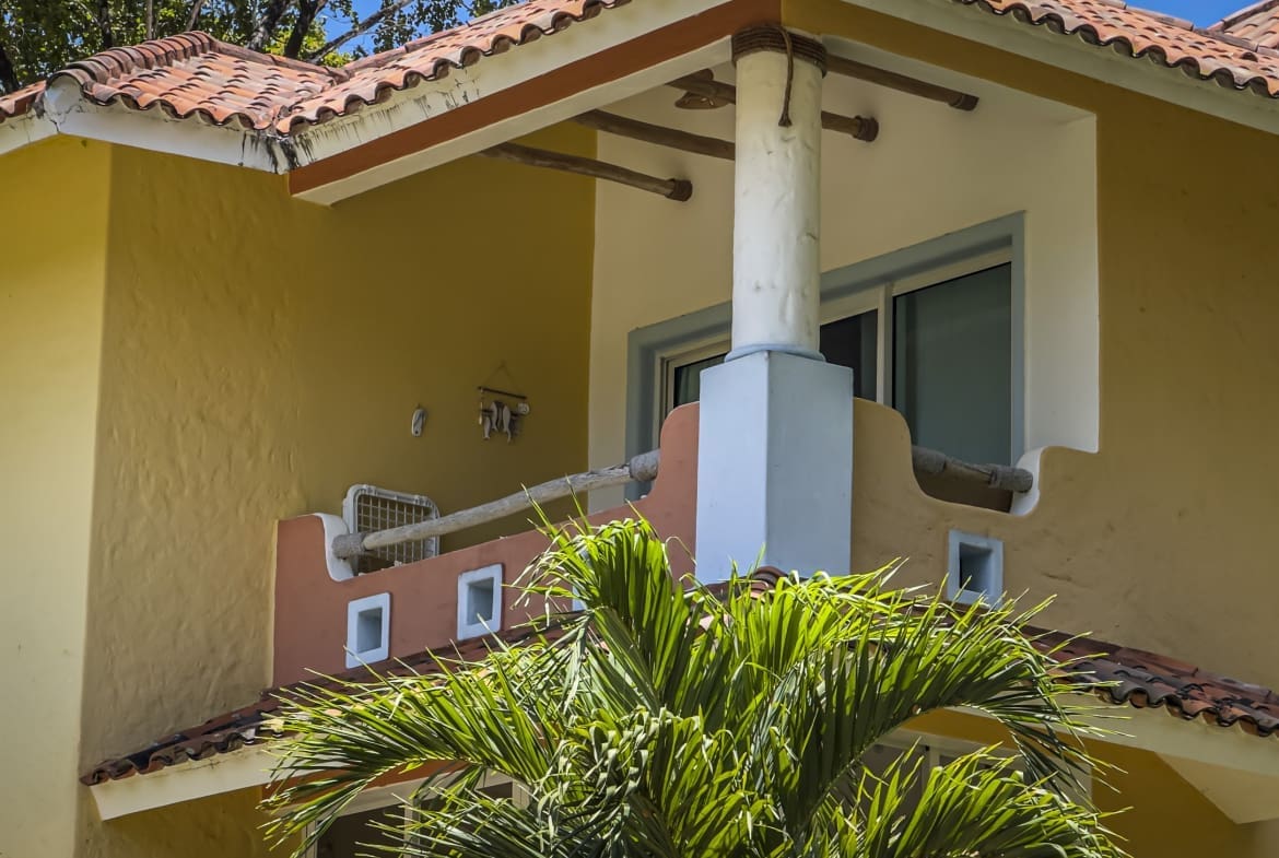 Yellow stucco house with red-tiled roof, white trim, and a small balcony framed by a white column; tropical plants in the foreground.