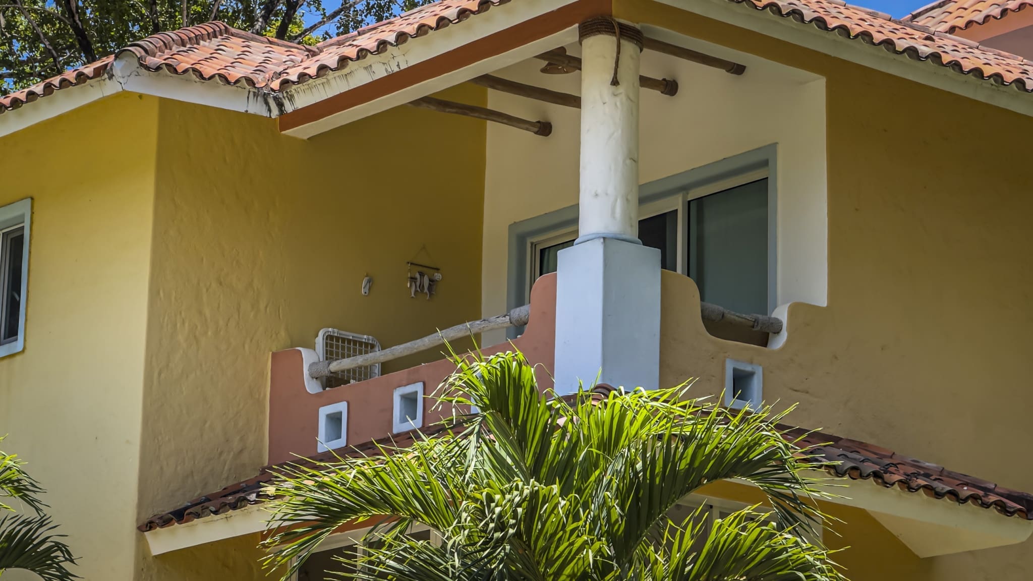 Yellow stucco house with red-tiled roof, white trim, and a small balcony framed by a white column; tropical plants in the foreground.