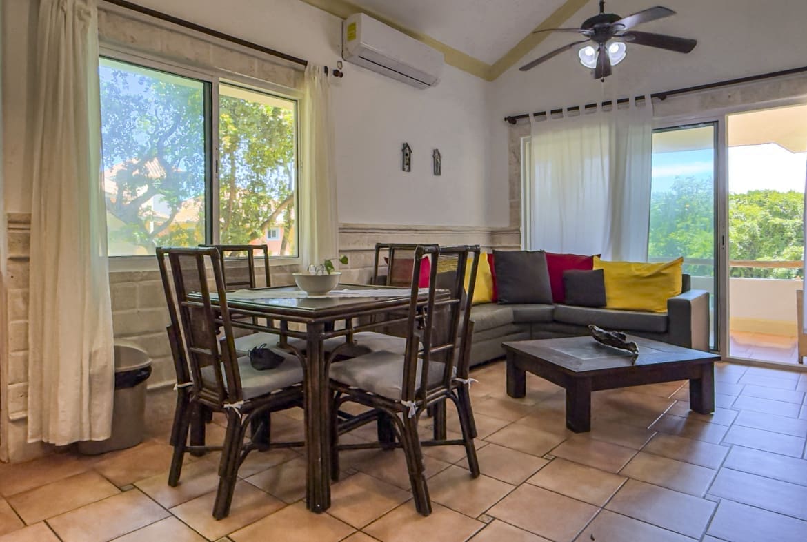 Living and dining area with a dark wood dining table and chairs, a gray sofa with colorful cushions, and large windows opening to a balcony.