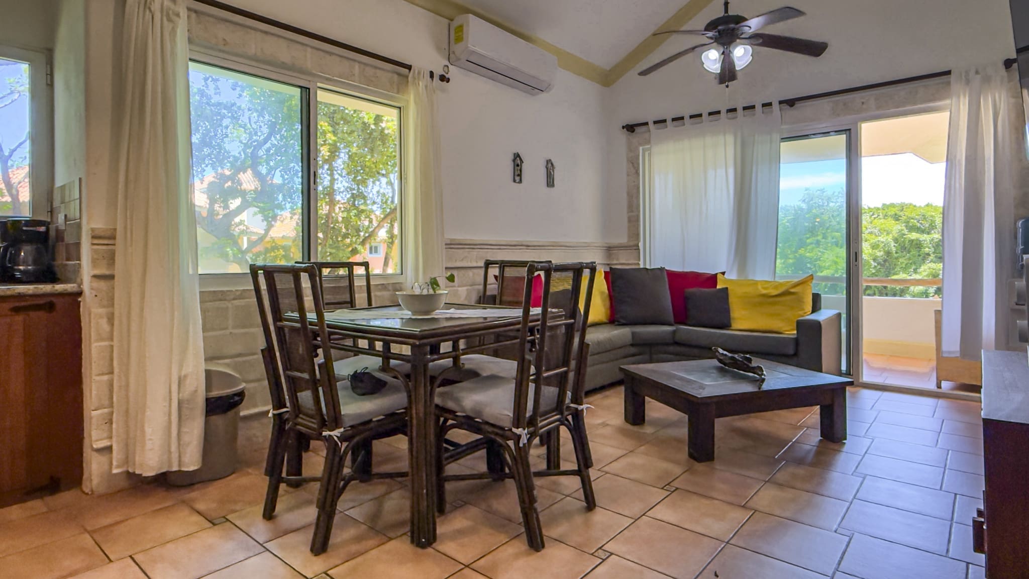Living and dining area with a dark wood dining table and chairs, a gray sofa with colorful cushions, and large windows opening to a balcony.