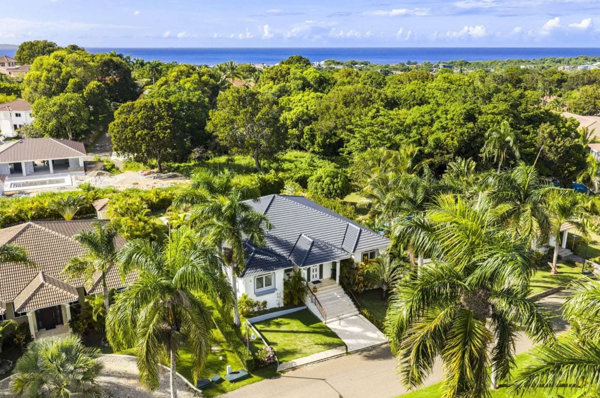 Aerial view of a white house with a gray roof surrounded by palm trees, with the ocean visible in the distance.