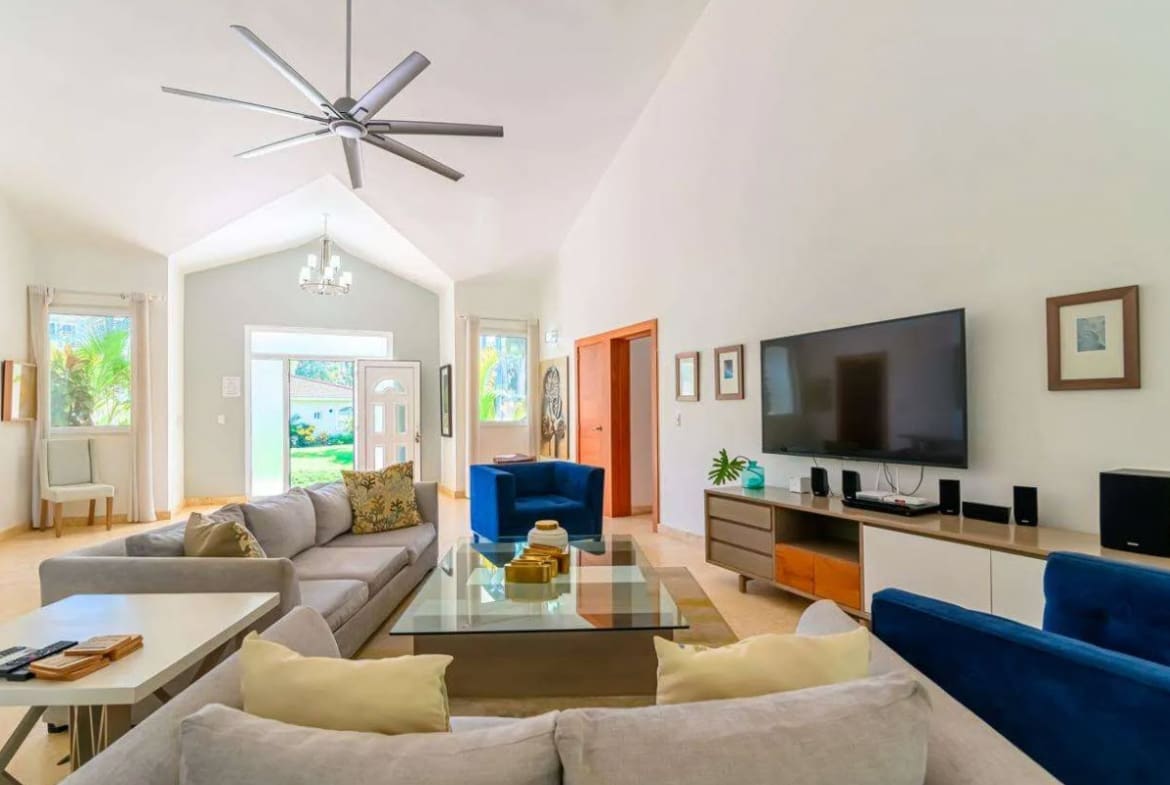 Bright open living room with a gray sectional, glass coffee table, and a wall-mounted TV in a sunlit space.