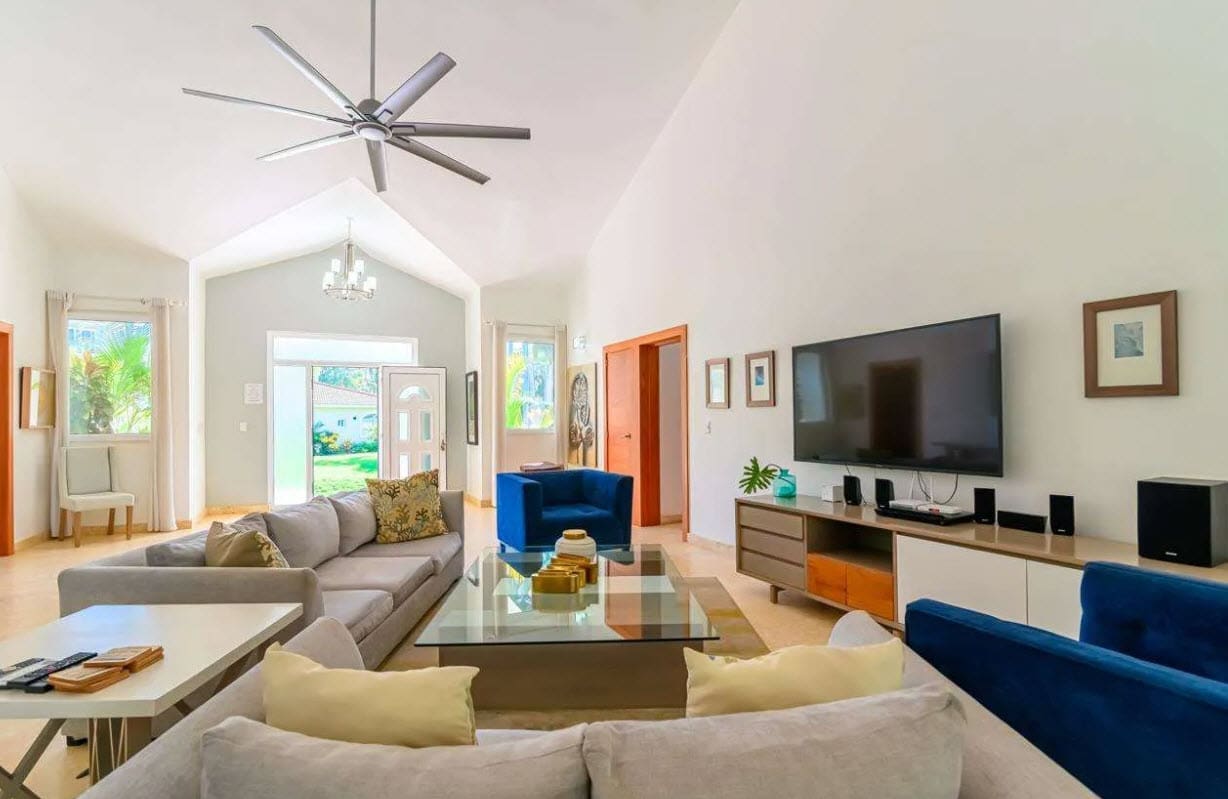Bright open living room with a gray sectional, glass coffee table, and a wall-mounted TV in a sunlit space.