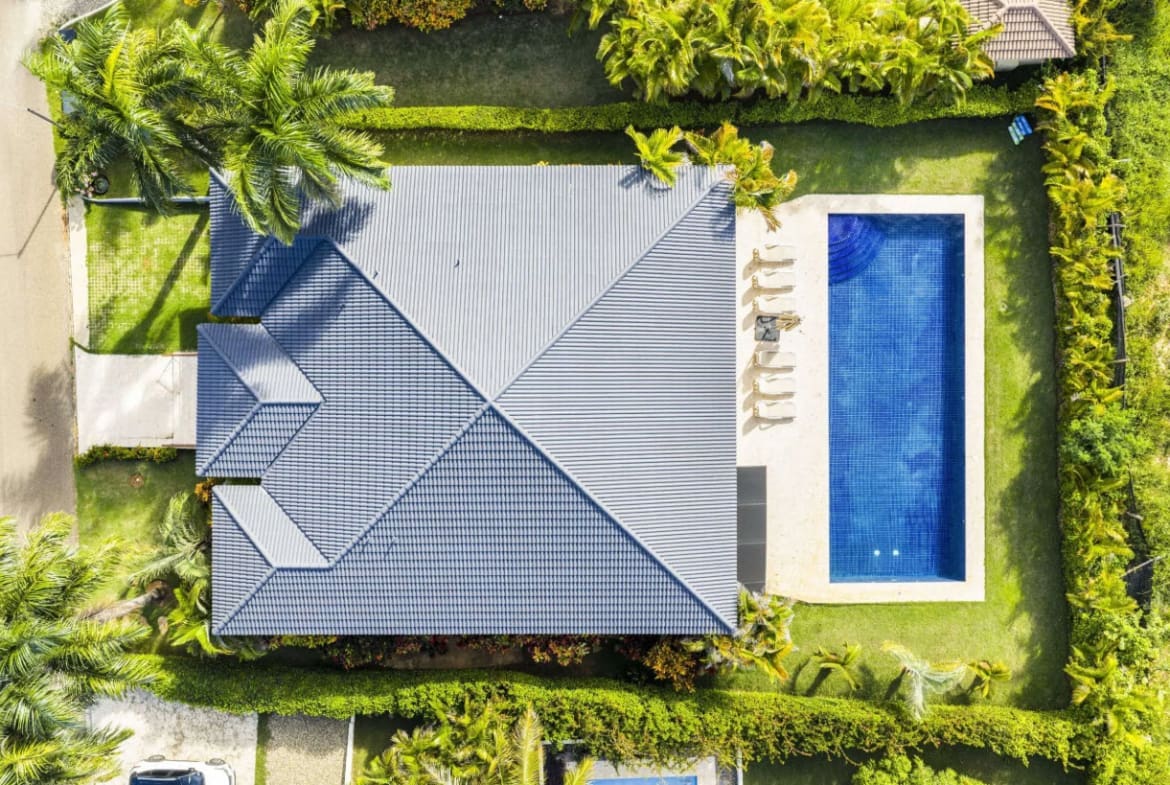Aerial view of a blue-tiled house with a rectangular blue swimming pool in a fenced yard, surrounded by green lawns and palm trees.