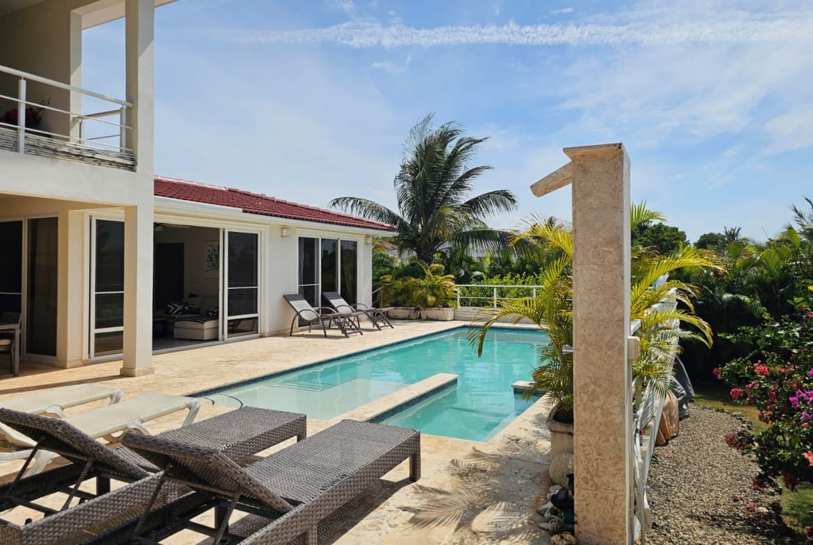 Resort-style backyard with a rectangular pool, beige stone patio, and white villa walls with sliding glass doors; lounge chairs nearby under a clear blue sky.