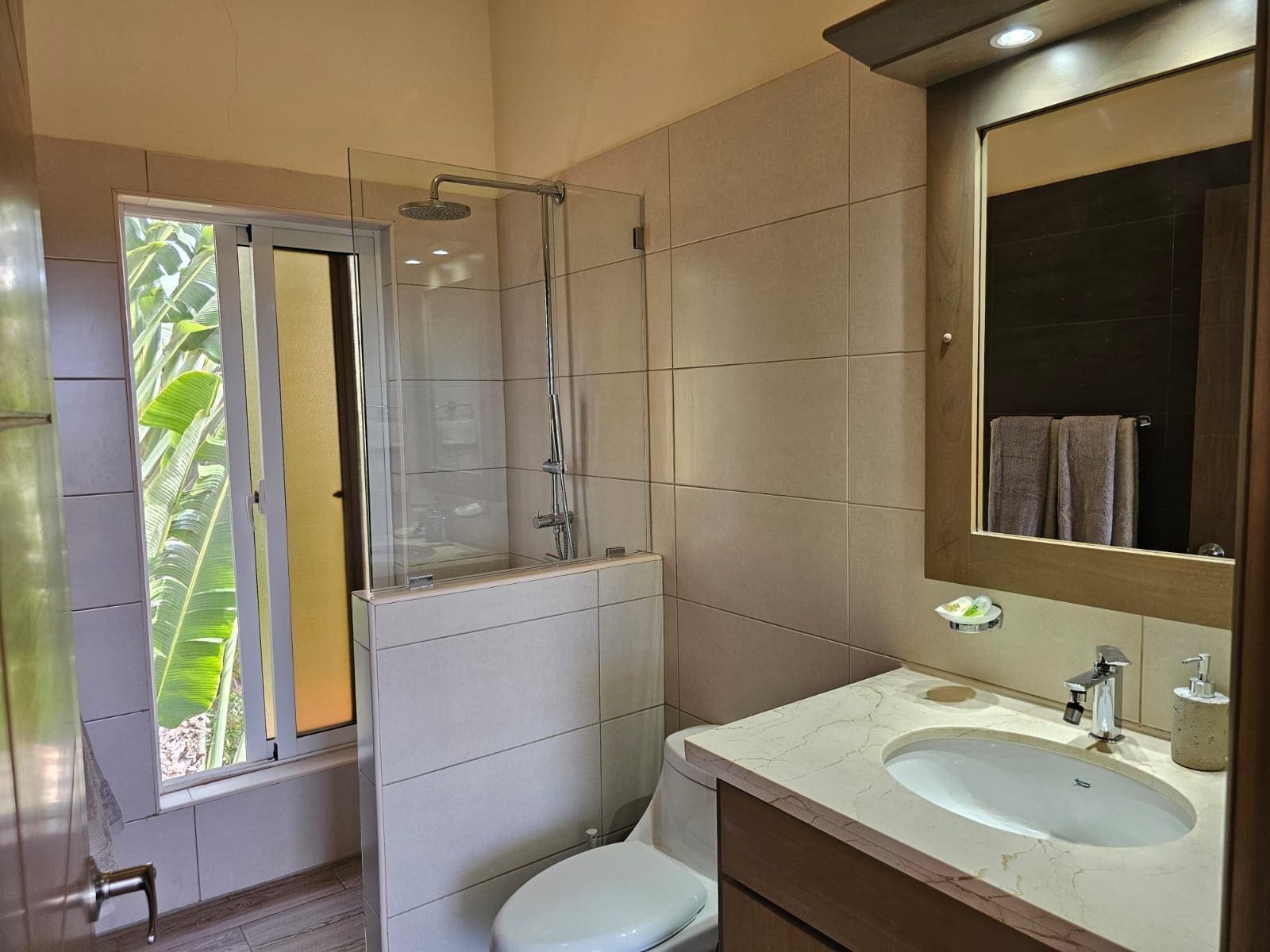 buyDRproperty Modern bathroom with walk-in shower, vanity with marble countertop, and large window with tropical foliage. Sosua Real Estate