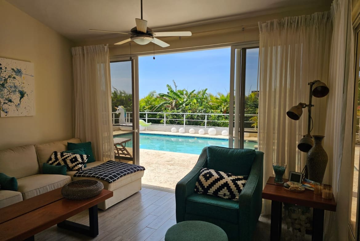 buyDRproperty Living room with teal armchair and sofa opens to a bright blue swimming pool and tropical greenery. Sosua Real Estate