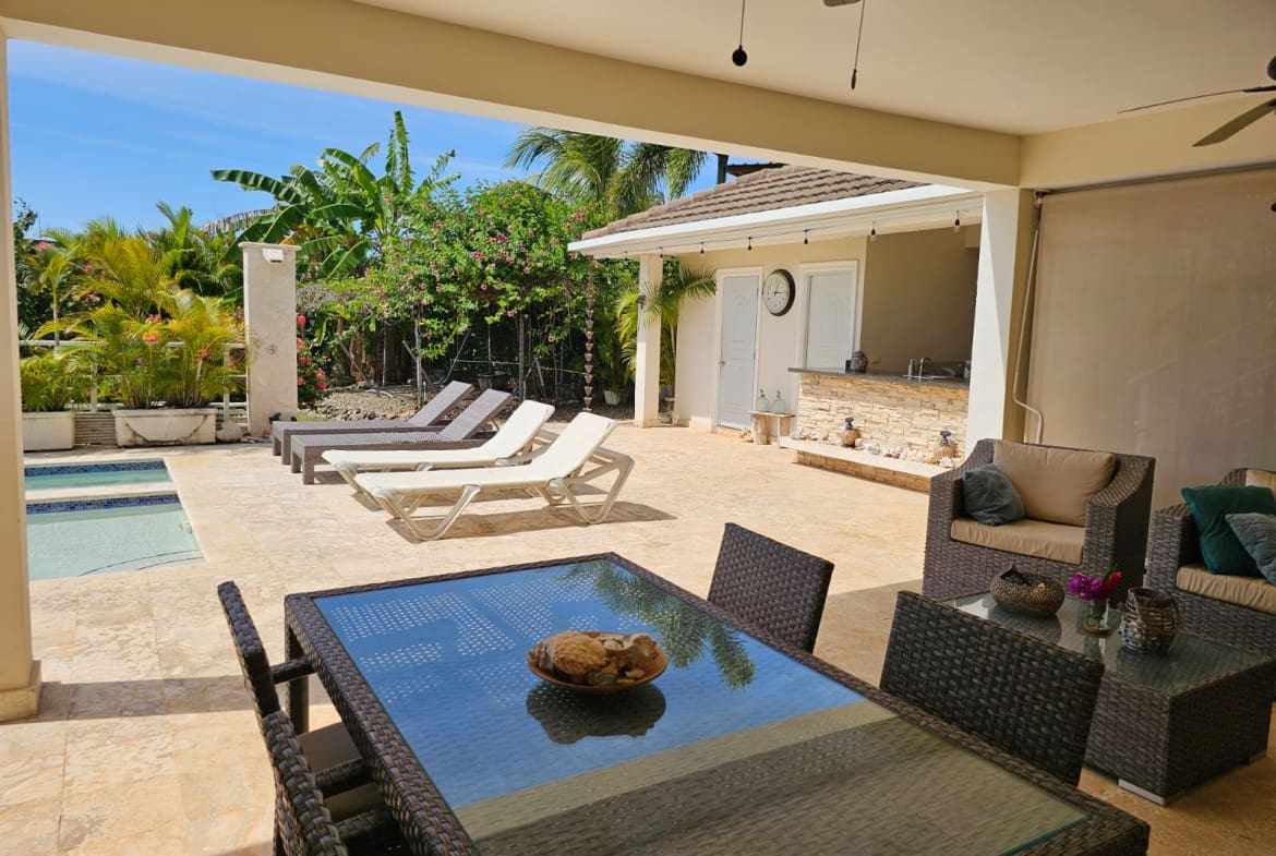 Outdoor pool patio with lounge chairs, wicker seating, and a bar under a shaded area amid tropical plants and palm trees.