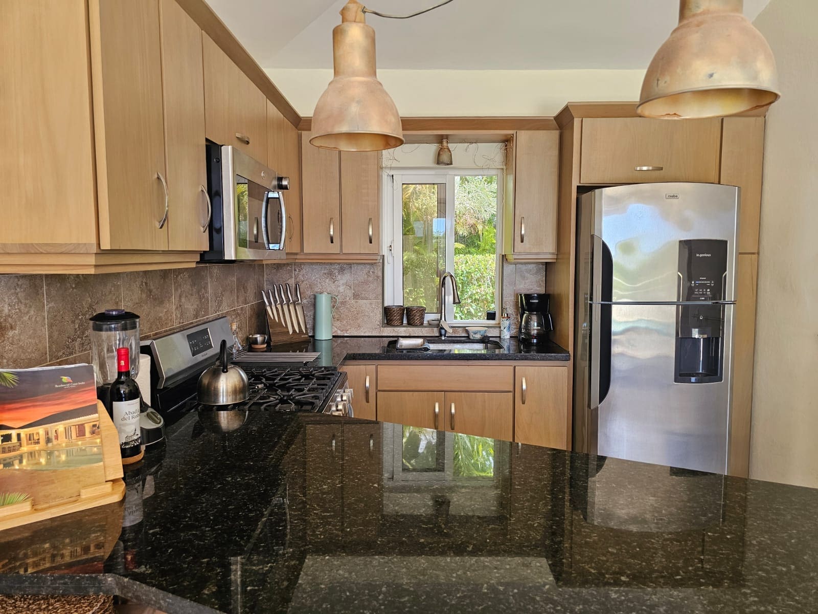 Modern L-shaped kitchen with wooden cabinets, black granite island, and stainless refrigerator under copper pendant lights. A window above the sink looks out to greenery.
