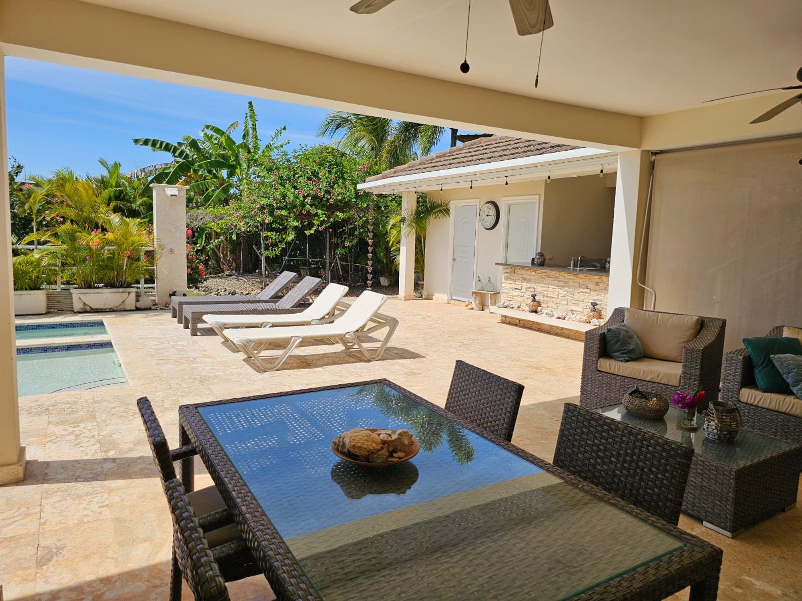 Outdoor pool patio with lounge chairs, wicker seating, and a bar under a shaded area amid tropical plants and palm trees.