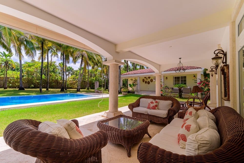 Covered patio with wicker sofas and a glass coffee table, opening to a blue pool and palm trees beyond.