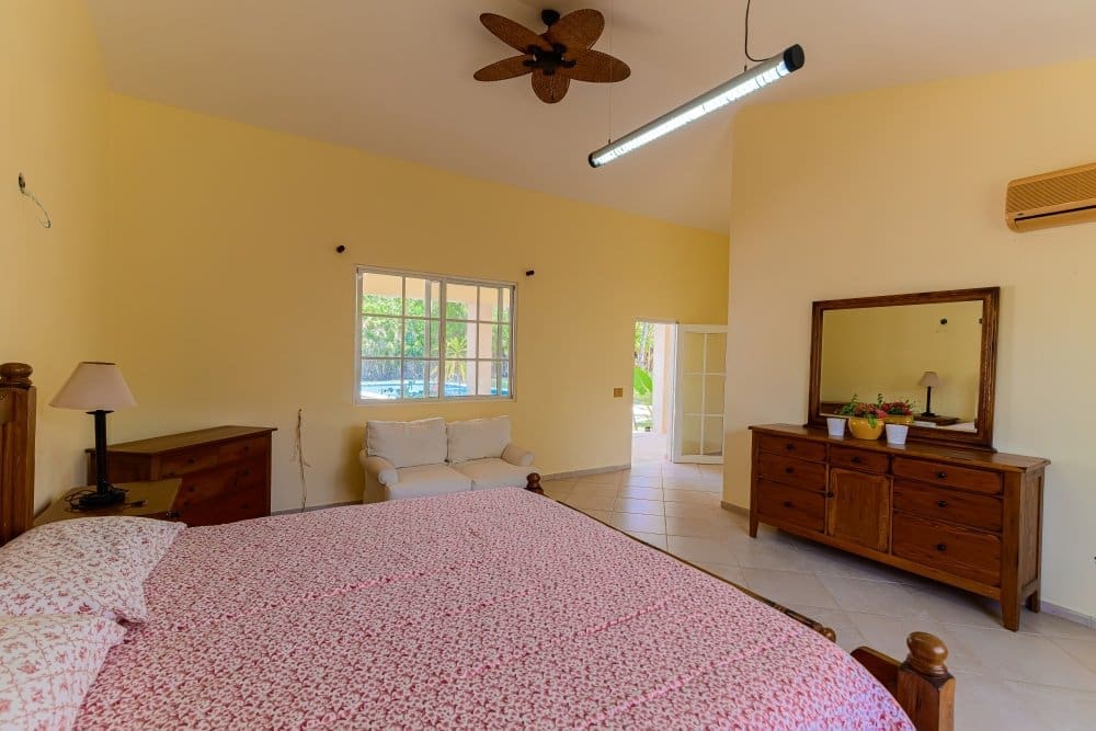 Sunlit bedroom with yellow walls, a large wooden bed covered in a pink floral quilt, and matching wooden dresser with a mirror. A small sofa sits by a window, and a ceiling fan hangs overhead.