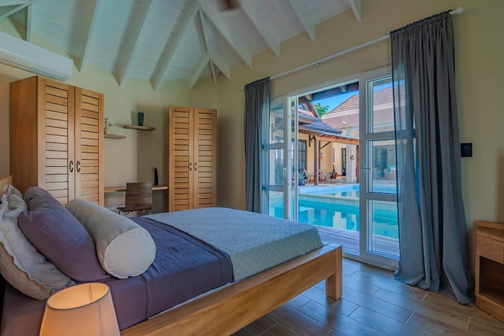 Bright bedroom with a wooden bed and blue bedding, opening to a poolside patio through large glass doors with blue curtains
