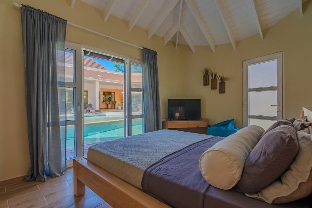 Bedroom with a wooden platform bed facing glass doors to a pool courtyard, TV on a dresser, and a vaulted white ceiling with exposed beams.