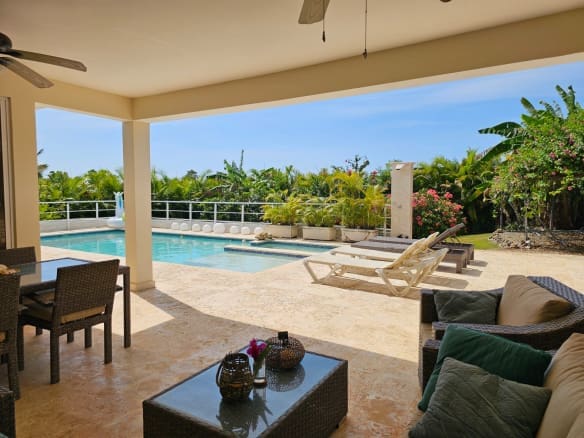 Covered patio area with wicker seating and a glass table overlooking a rectangular pool and tropical garden.