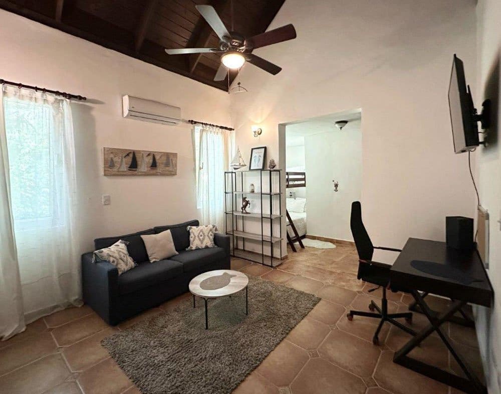 buyDRproperty Cozy living room with a dark gray sofa, rug, and desk, featuring a ceiling fan and natural light. Sosua Real Estate