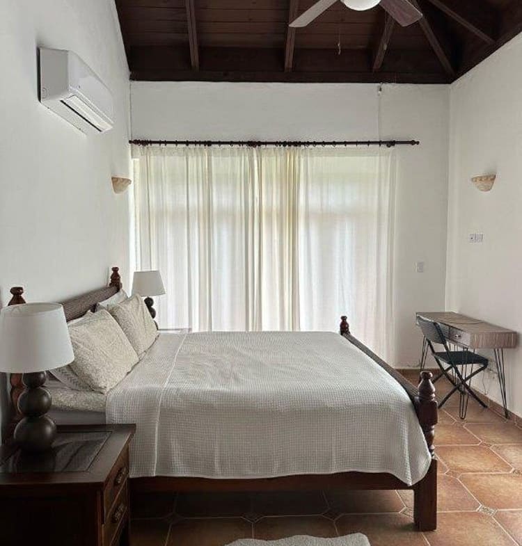 Bedroom with white bedding and dark wooden bed frame, two nightstands, and a ceiling fan; sheer curtains over glass doors in back of room.
