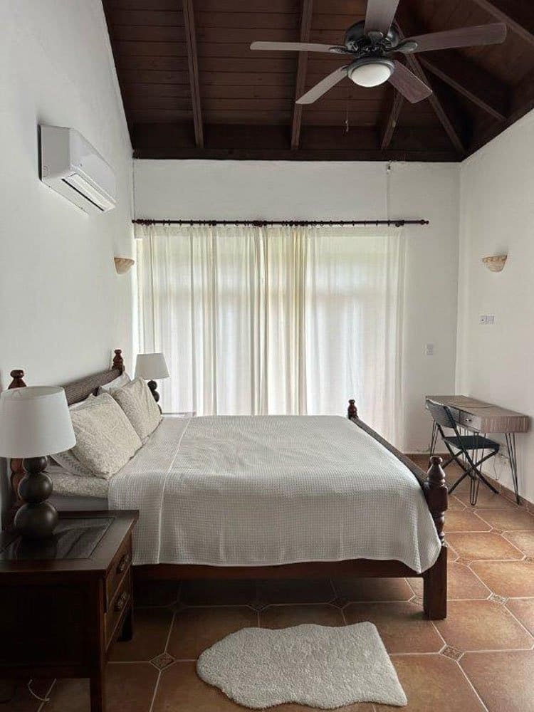 Bedroom with white bedding and dark wooden bed frame, two nightstands, and a ceiling fan; sheer curtains over glass doors in back of room.
