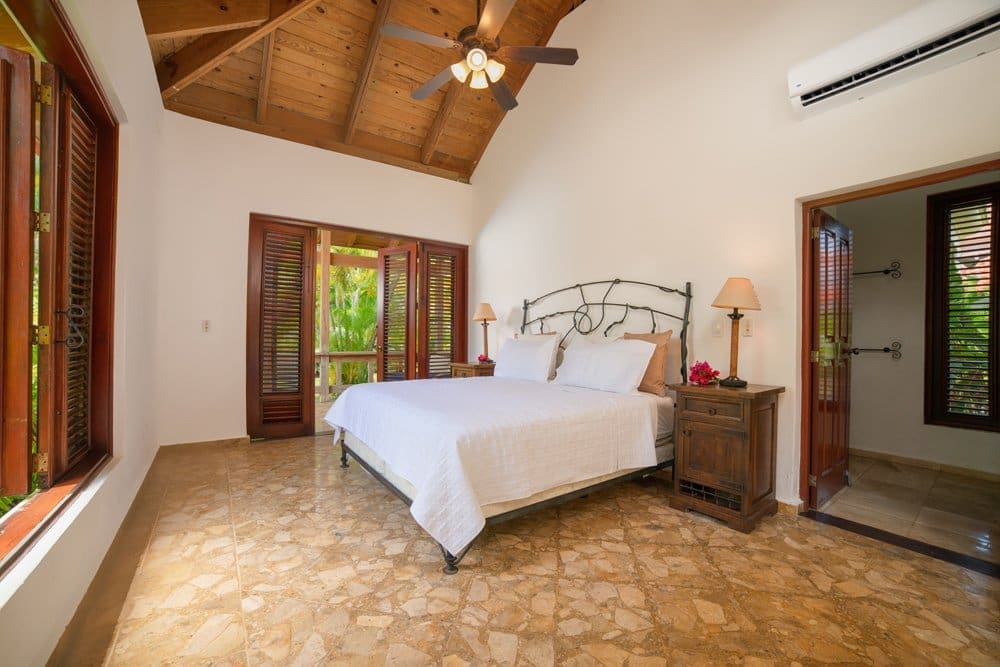 buyDRproperty Tropical bedroom with white bedding, wooden shutters, and AC unit. Sosua Real Estate