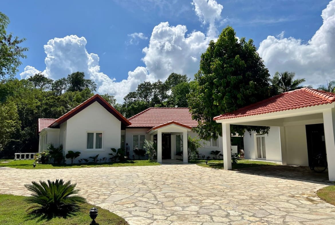 buyDRproperty White modern house with red tile roof and stone driveway in lush green landscape. Sosua Real Estate